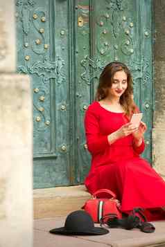 美丽的年轻的高加索人女人坐着楼梯通过旅行手提箱智能手机穿长红色的衣服