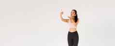 完整的长度可爱的愚蠢的女博主健身房穿运动服装采取自拍生活流健身房锻炼会话发送空气吻移动电话屏幕站白色背景