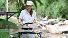 快乐女人旅行者女人烧烤肉可移植的气体炉子野营河银行冒险野营概念