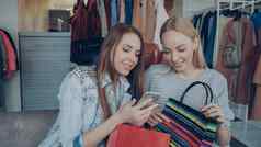 有吸引力的年轻的女士们坐着服装商店咖啡购物袋检查智能手机会说话的货架上衣架衣服背景