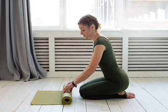 女人绿色连身裤曲折绿色橡胶席健身瑜伽坐着明亮的房间窗口