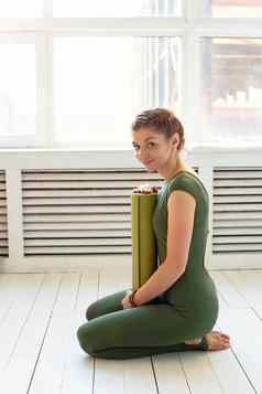 纤细的美丽的女人绿色体育连身裤坐在持有扭曲的绿色席健身瑜伽