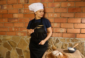 漂亮的女人手工家庭面包店老板贝克酋长帽围裙摆姿势小麦耳朵砖墙