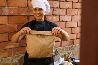 美丽的女人面包店商店工人持有纸袋包装新鲜的传统的面包可爱的微笑相机
