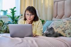 中间岁的女人放松首页床上移动PC宠物猫
