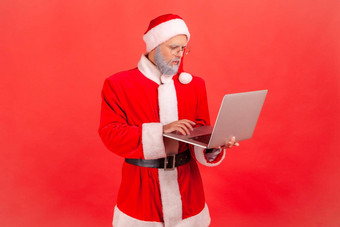 上了年纪的男人。灰色的胡子穿圣诞老人老人服装工作电脑显示集中面部表达式