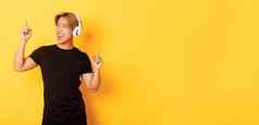 快乐的英俊的亚洲的家伙金发碧眼的头发唱歌跳舞听音乐无线耳机站黄色的背景