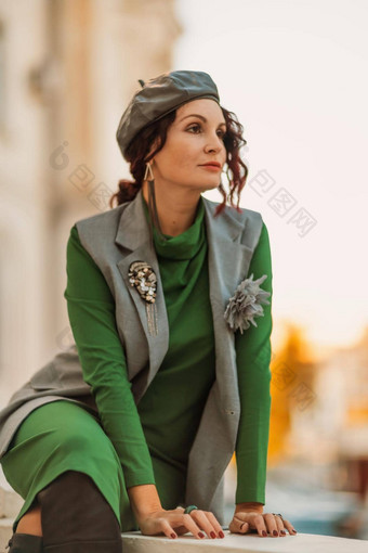 户外时尚肖像优雅的时尚浅黑肤色的女人模型贝雷帽绿色衣服灰色的马甲摆姿势日落欧洲城市秋天