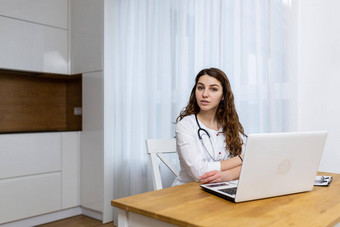 年轻的女医生<strong>对待</strong>在线首页咨询视频调用移动PC坐着厨房白色外套相机