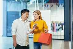 多样化的夫妇亚洲男人。金发女郎女人衣服超市移动电话屏幕选择购物持有色彩斑斓的购物袋礼物