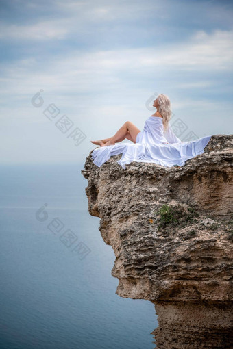 金发女郎坐在岩石海长头发阳光明媚的海滨白色衣服后视图丝绸织物收敛了风背景蓝色的天空山