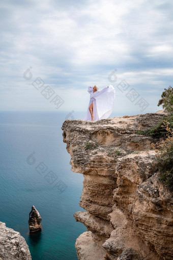 金发女郎长头发阳光明媚的海滨白色流动衣服后视图丝绸织物挥舞着风背景蓝色的天空山海滨