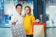 年轻的多样化的家庭亚洲男人。金发女郎女人购物超市肖像夫妇购物者相机微笑持有色彩斑斓的袋货物购物