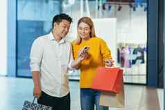 多样化的夫妇亚洲男人。金发女郎女人衣服超市移动电话屏幕选择购物持有色彩斑斓的购物袋礼物