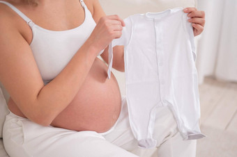 怀孕了女人坐在白色沙发持有衣服<strong>预期</strong>婴儿