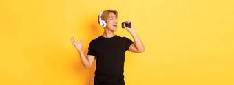 快乐的有吸引力的朝鲜文的家伙耳机玩<strong>卡拉ok</strong>应用程序唱歌移动电话麦克风站黄色的背景
