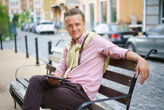 自由职业者年轻的男人。数字平板电脑手坐在板凳上在户外前面办公室建筑穿休闲粉红色的衬衫肩膀袋年轻的英俊的男人。工作