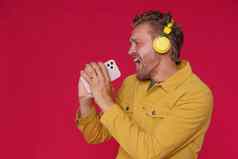 英俊的男人。唱歌享受最喜欢的首歌跟踪电话无线耳机穿牛仔布黄色的夹克孤立的红色的背景快乐的男人。唱听音乐