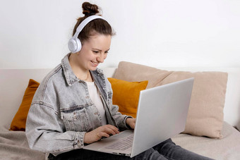年轻的高加索人女人耳机坐着床上持有移动PC电脑女孩笔记本冲浪互联网读新闻看电影研究工作在线调用朋友放松首页