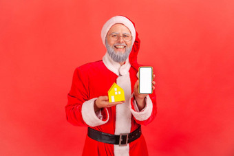 肖像微笑上了年纪的男人。灰色的胡子穿圣诞老人老人服装持有小纸房子聪明的电话白空白屏幕