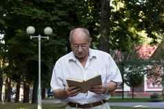 上了年纪的退休男人。走公园秃顶男人。眼镜看手站背景红色的建筑读取书