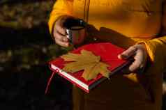 女人持有热水瓶热茶红色的书黄色的下降枫木叶秋天