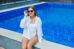 快乐的年轻的女人白色泳衣白色衬衫坐着池女人太阳镜泳衣夏天假期池晒黑女孩