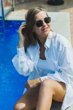 快乐的年轻的女人白色泳衣白色衬衫坐着池女人太阳镜泳衣夏天假期池晒黑女孩