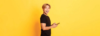 肖像英俊的时尚的亚洲的家伙黑色的装移动电话把头相机满意微笑黄色的背景
