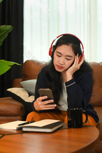 漂亮的年轻的亚洲女人穿无线耳机移动电话沙发上首页