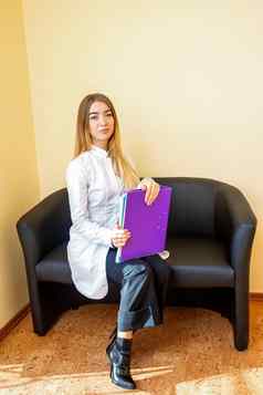 肖像年轻的高加索人医生文件夹金发女郎长头发穿白色外套坐着黑色的沙发相机