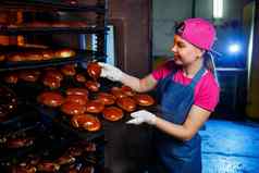 年轻的女孩贝克持有托盘热糕点背景工业烤箱面包店生产面包店产品新鲜的脆皮糕点架