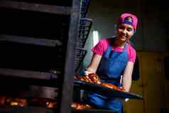 年轻的女孩贝克持有托盘热糕点背景工业烤箱面包店生产面包店产品