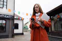 年轻的女人秋天橙色外套移动PC移动电话背景城市