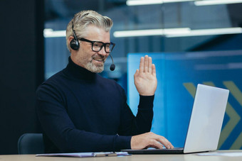 在线会议的同事们高级头发花白的男人。工作现代办公室室内执行耳机眼镜微笑移动PC相机挥舞着问候手势视频调用