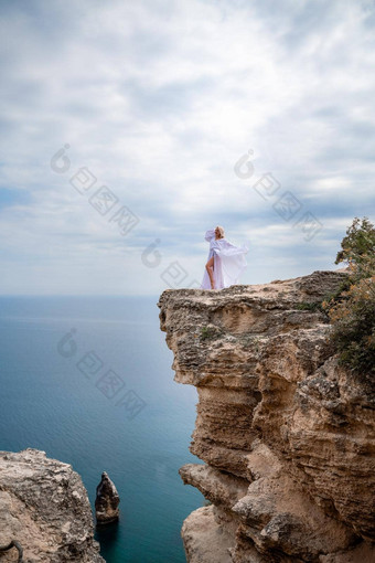 金发女郎长头发阳光明媚的海滨白色流动衣服后视图丝绸织物挥舞着风背景蓝色的天空山海滨