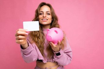 照片拍摄年轻的美丽的有吸引力的可爱的积极的快乐微笑卷曲的金发女郎女人穿时尚的时尚的粉红色的体育运动西装孤立的粉红色的背景复制空间文本持有粉红色的一分钱猪白色银行塑料卡钱
