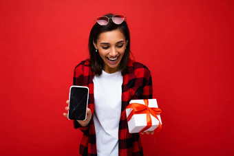 有吸引力的快乐年轻的浅黑肤色的女人女人孤立的红色的背景墙穿白色休闲t恤红色的黑色的衬衫持有白色礼物盒子红色的丝带移动电话空屏幕显示模型