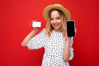 拍摄美丽的快乐微笑金发女郎女人夏天衣服显示智能手机空屏幕模拟信贷卡孤立的红色的背景业务概念