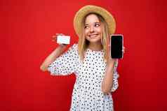 拍摄美丽的快乐微笑金发女郎女人夏天衣服显示智能手机空屏幕模拟信贷卡孤立的红色的背景业务概念