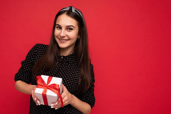 拍摄迷人的快乐微笑浅黑肤色的女人女孩孤立的红色的背景墙穿黑色的上衣持有白色礼物盒子红色的丝带相机