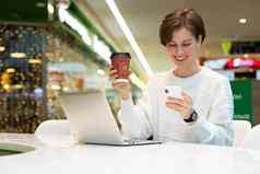 年轻的美丽的女人坐着购物中心表格工作电脑移动PC移动电话喝咖啡自由业务概念
