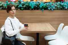 年轻的美丽的女人穿白色运动衫坐着购物中心表格喝咖啡