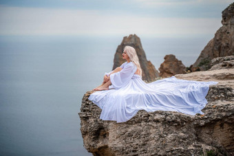 金发女郎坐在岩石海长头发阳光明媚的海滨白色衣服后视图丝绸织物收敛了风背景蓝色的天<strong>空山</strong>