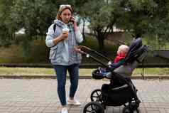 年轻的女人走婴儿婴儿车会说话的移动电话喝咖啡城市公园