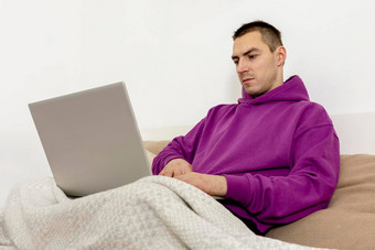 年轻的高加索人男人。紫罗兰色的连帽衫坐着床上持有移动<strong>PC</strong>电脑男人。笔记本冲浪互联网读<strong>新</strong>闻看电影研究工作在线放松<strong>首页</strong>