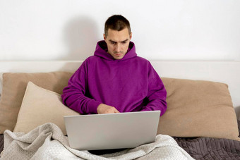 年轻的高加索人男人。紫罗兰色的连帽衫坐着床上持有移动PC电脑男人。笔记本冲浪互联网读新闻看电影研究工作在线放松首页