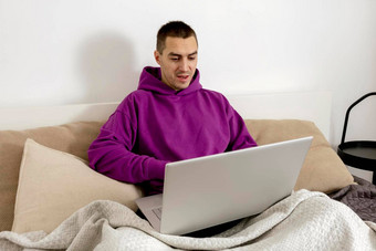 年轻的高加索人男人。紫罗兰色的连帽衫坐着床上持有移动PC电脑男人。笔记本冲浪互联网读新闻看电影研究工作在线调用朋友放松首页