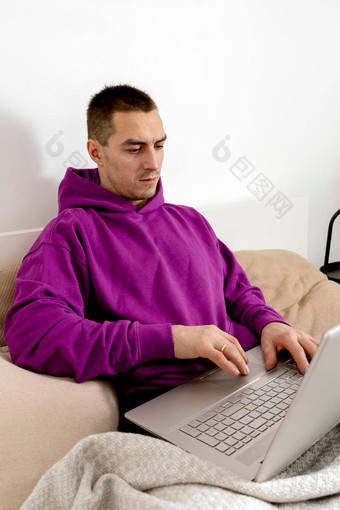 年轻的高加索人男人。紫罗兰色的连帽衫坐着床上持有移动PC电脑男人。笔记本冲浪互联网读新闻看电影研究工作在线调用朋友放松首页