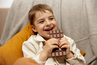 可爱的男孩坐着沙发上<strong>首页</strong>吃巧克力酒吧孩子糖果糖糖果孩子享受美味的<strong>甜点</strong>学前教育孩子休闲服装积极的情感